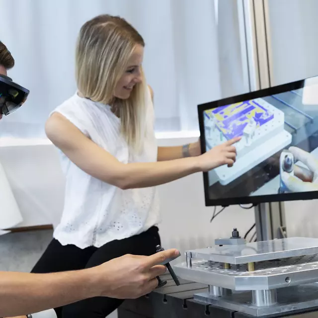 Zwei Studierende der Fakultät Maschinenbau an der OTH Regensburg arbeiten im Labor Fertigungstechnik und Werkzeugmaschinen. Der männliche Student trägt eine VR-Brille und führt eine Tätigkeit mit einem Controller durch. Die weibliche Studentin deutet auf einen Computerbildschirm, der die Handlung des Studenten visualisiert.