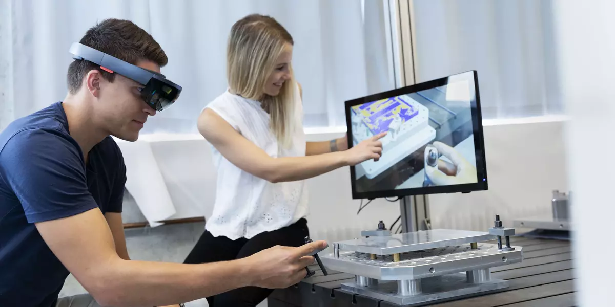 Zwei Studierende der Fakultät Maschinenbau an der OTH Regensburg arbeiten im Labor Fertigungstechnik und Werkzeugmaschinen. Der männliche Student trägt eine VR-Brille und führt eine Tätigkeit mit einem Controller durch. Die weibliche Studentin deutet auf einen Computerbildschirm, der die Handlung des Studenten visualisiert.